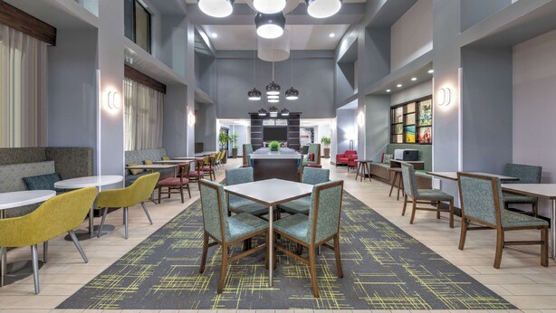 A modern dining area with tables and chairs, a central seating area, and a kitchen in the background. - Hampton Inn & Suites Lake Jackson-Clute (Clute)