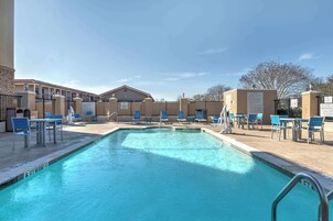 Zona de piscina con sillas y mesas azules, cielo despejado y un edificio al fondo.