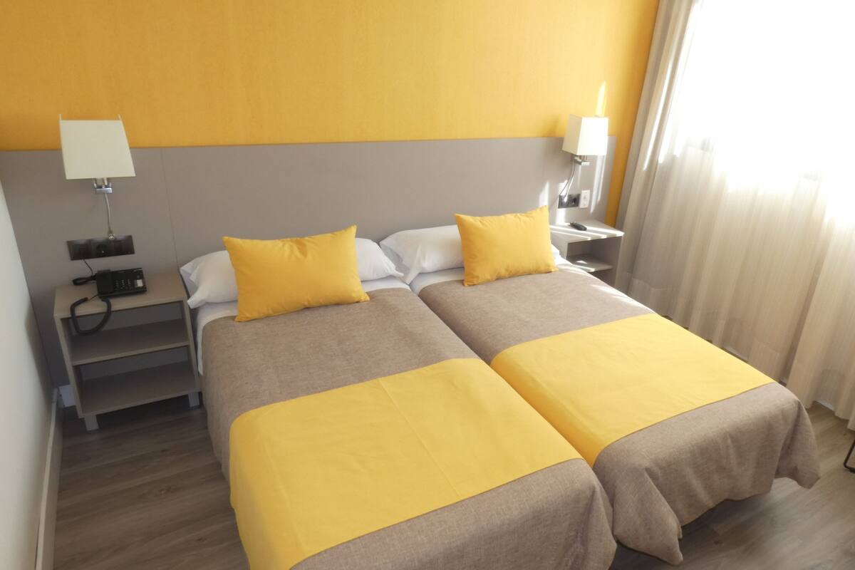 Ein Hotelzimmer mit zwei Betten, einem Schreibtisch mit Telefon, einem Sessel und einem Fenster mit Vorhängen.