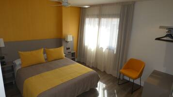 Habitación de hotel con cama, una silla, escritorio, lámpara, mesita de noche y ventana con cortinas.