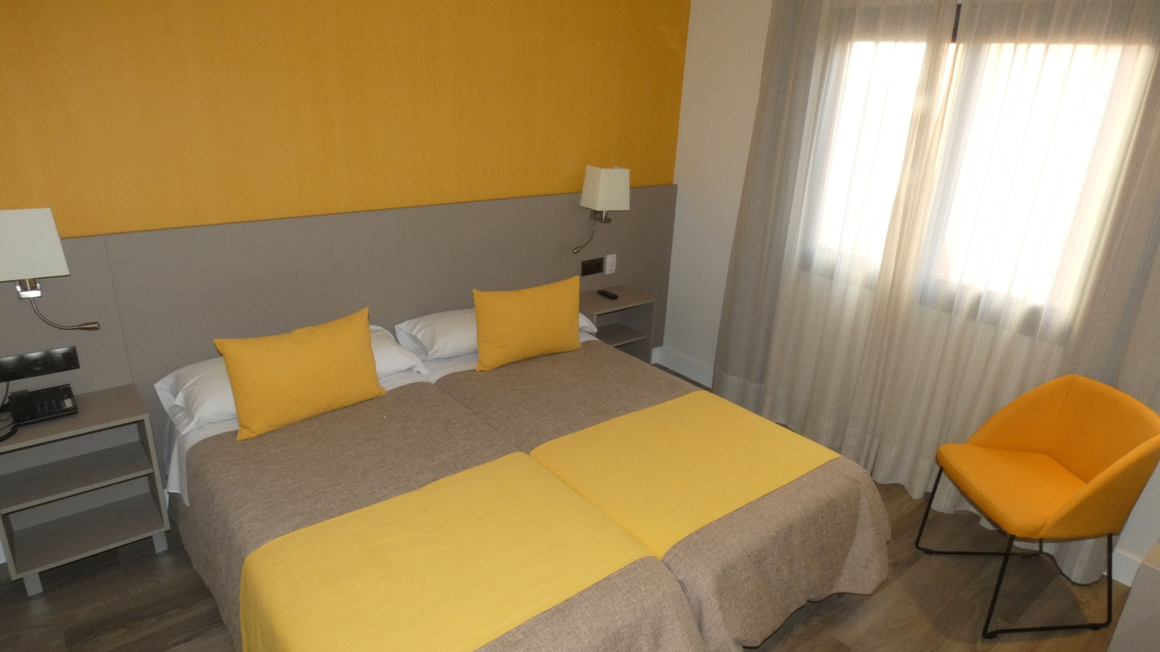 A hotel room with a double bed, two pillows, a bedside table with a lamp, a chair, and a window with curtains.