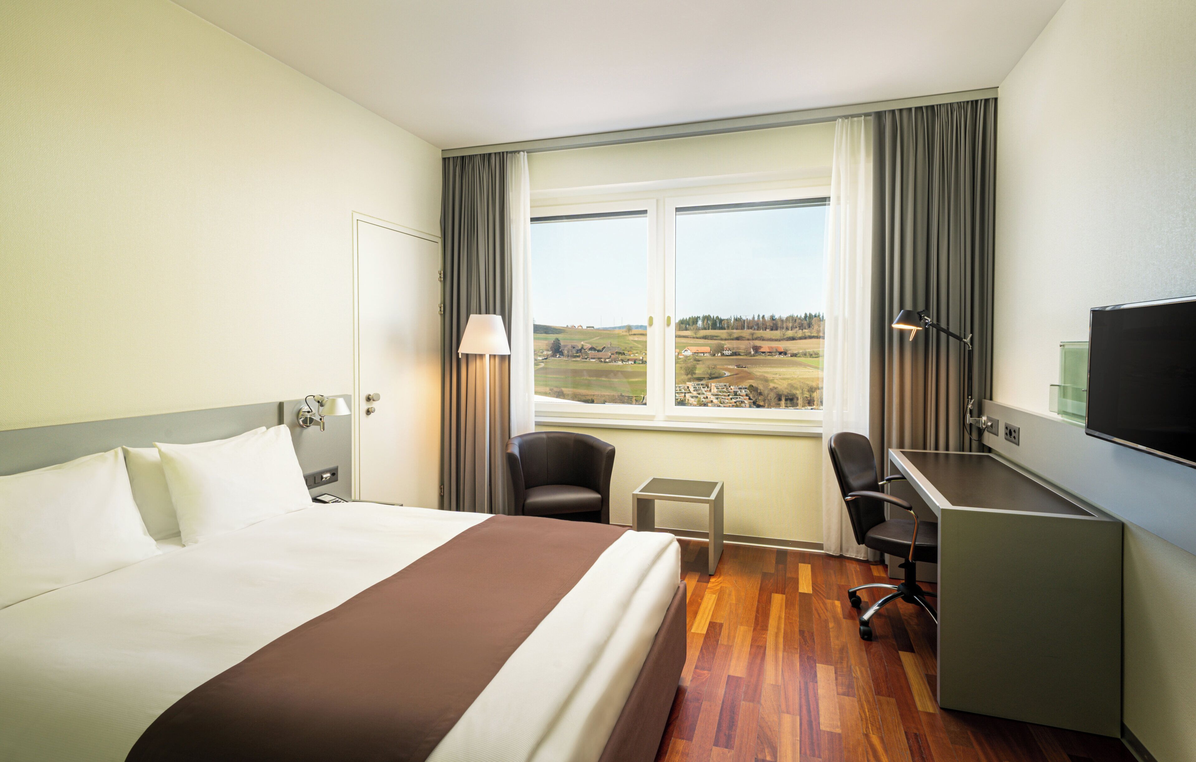 Ein Hotelzimmer mit Bett, Schreibtisch, Stuhl, Fernseher und einem Fenster mit Blick ins Grüne.