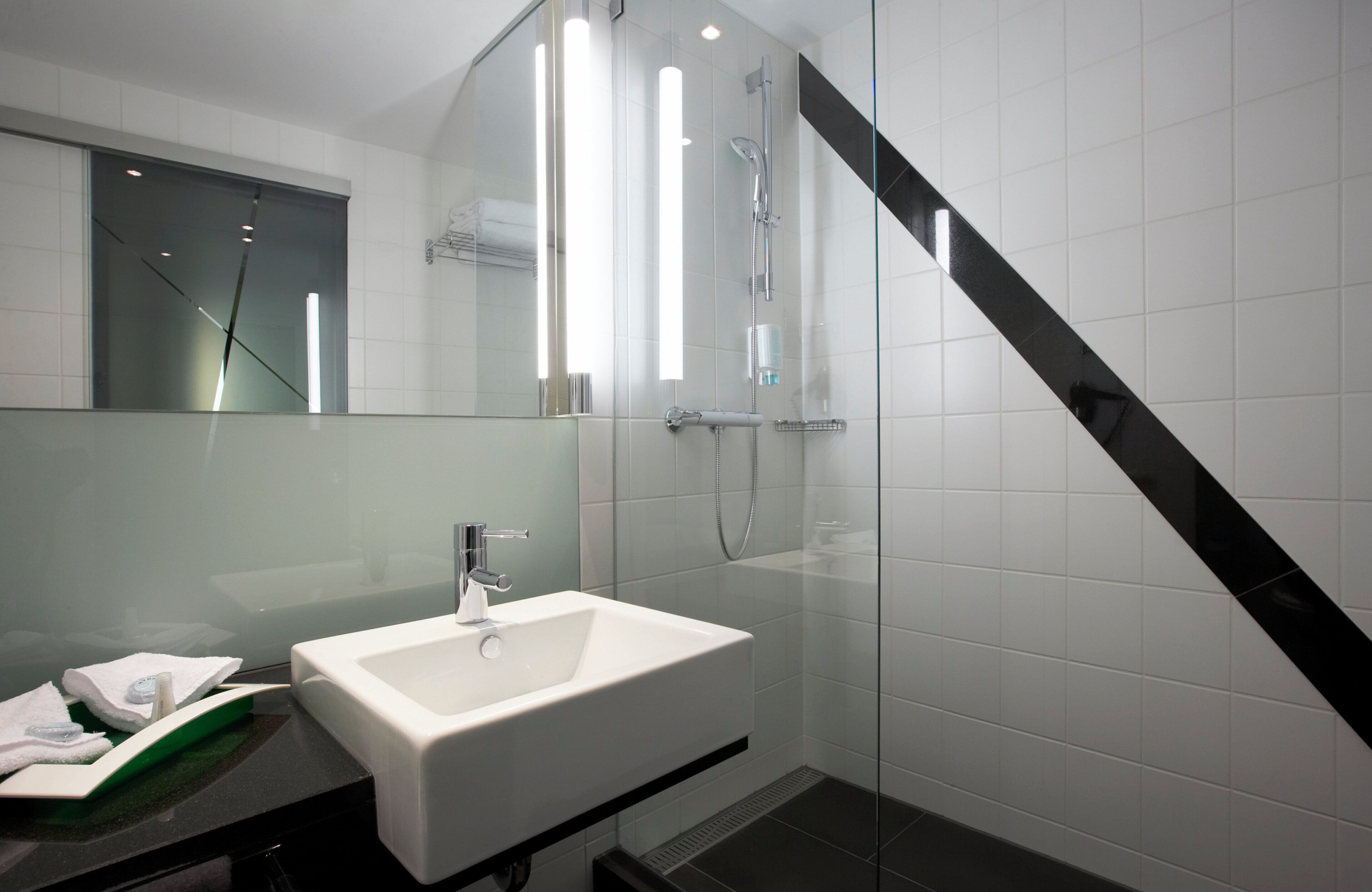 Ein modernes Badezimmer mit Waschbecken in Weiß, einer Duschkabine aus Glas und einem schwarzen, diagonalen Akzent an der Wand.