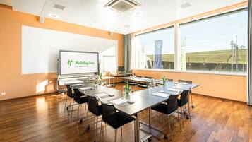 Un salón de conferencias con una gran mesa, sillas, una pantalla de proyector mostrando 'Holiday Inn EIN IHG HOTEL' y vistas a un campo a través de amplias ventanas.