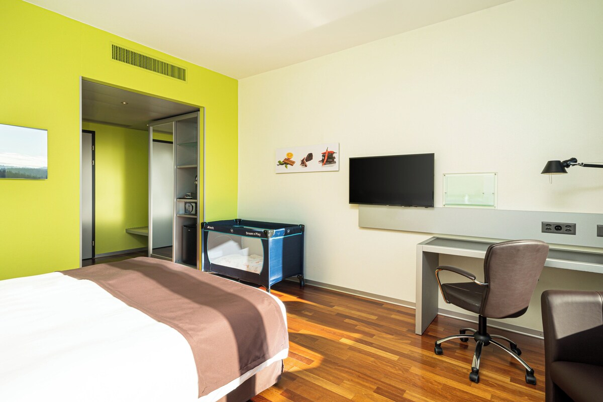 A hotel room with a bed, a desk, a chair, a television, and a small playpen.