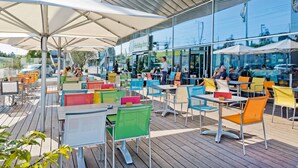 An outdoor cafe area with tables and chairs, large umbrellas, and a glass building in the background. - Holiday Inn BERN-WESTSIDE by IHG (Bern)