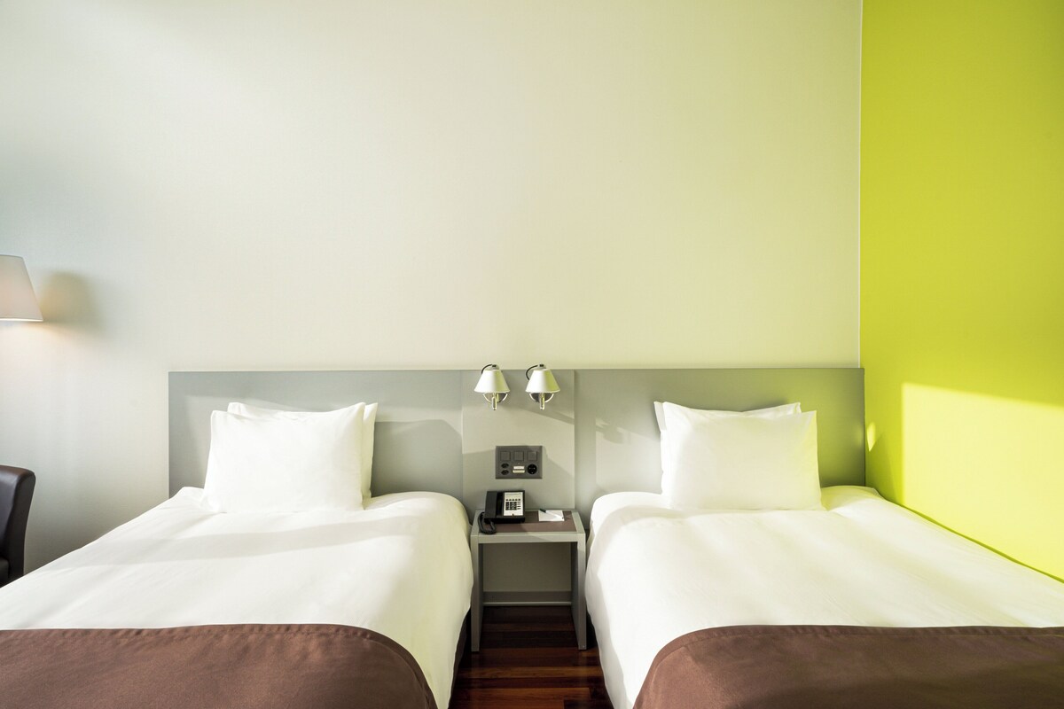 A hotel room with two beds, a nightstand with a phone and lamps, and a chair.