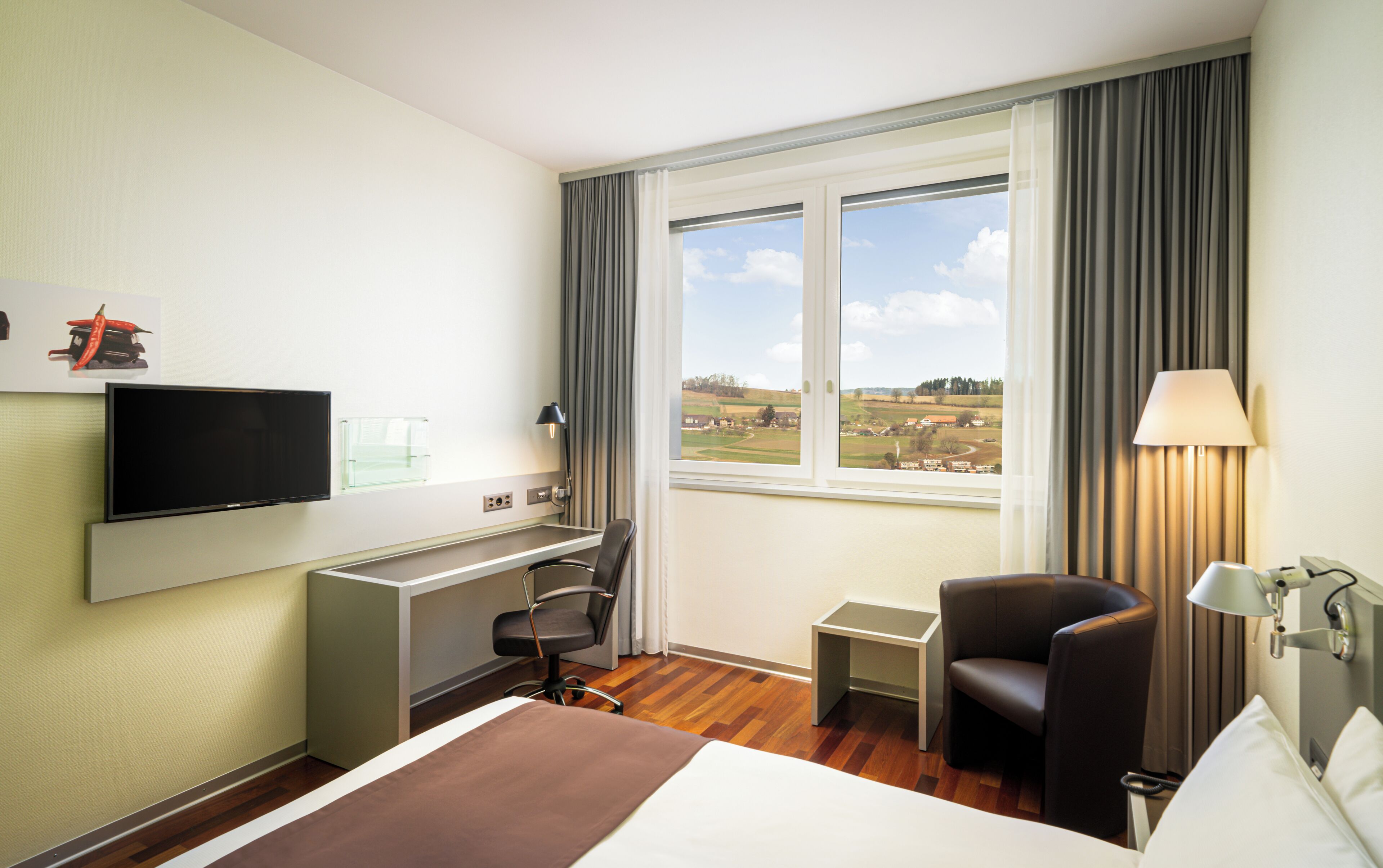 Habitación de hotel con cama, escritorio, silla, televisión, lámpara y ventana con vistas al campo.