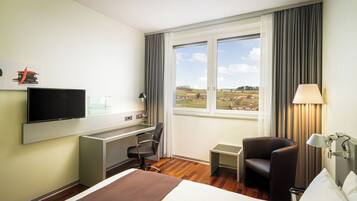 Habitación de hotel con cama, escritorio, silla, televisión, lámpara y ventana con vistas al campo.