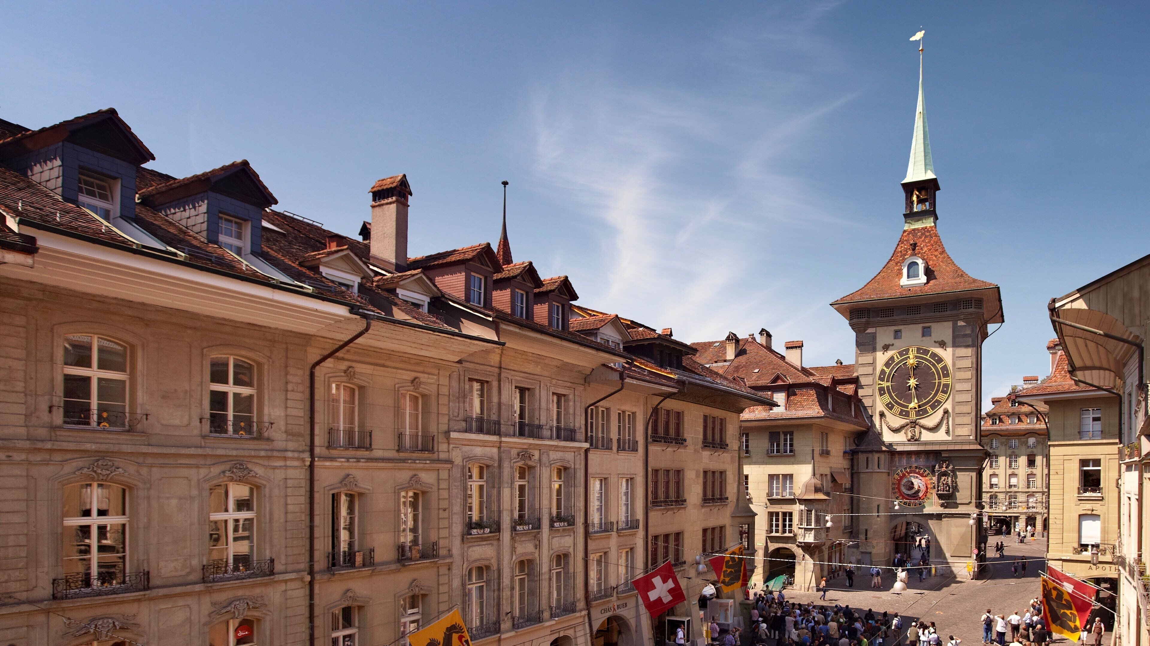 A historic building with a prominent clock tower, a traditional facade, and a public square with flags.
