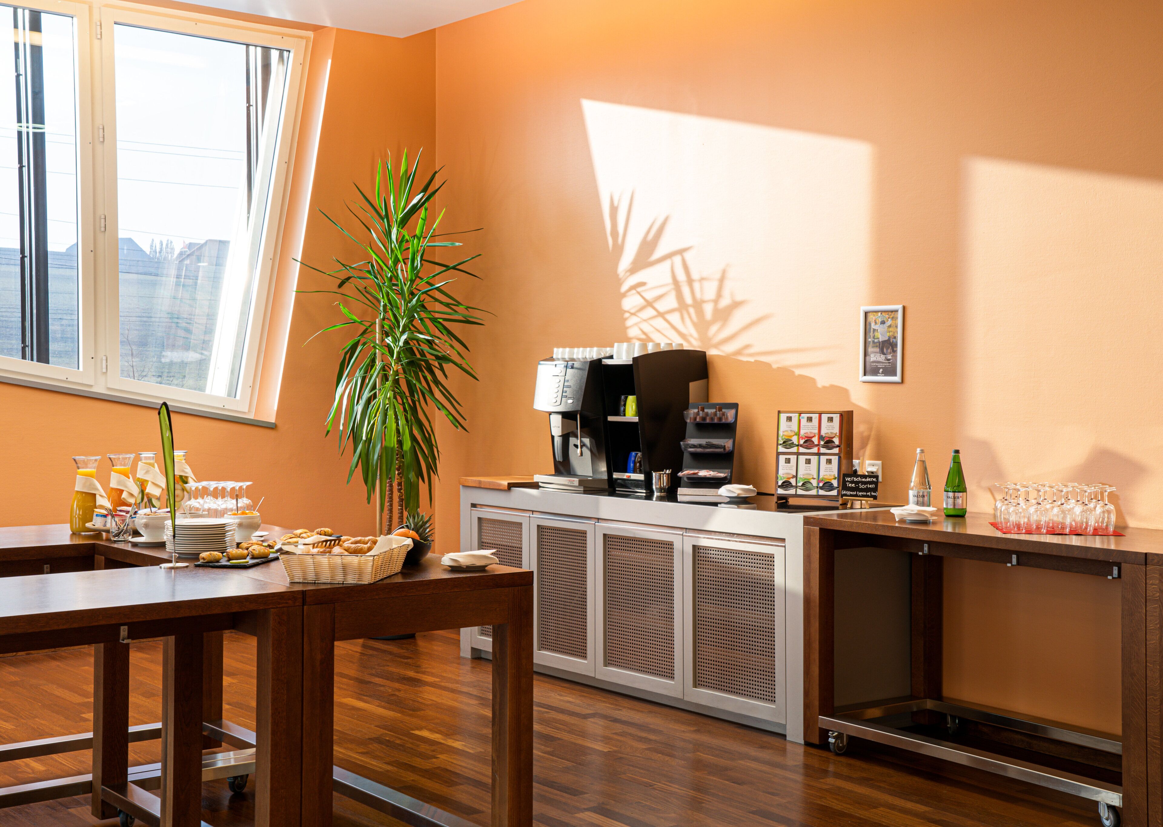 A hotel room with a dining area, a coffee station, a plant, and a view of the city.