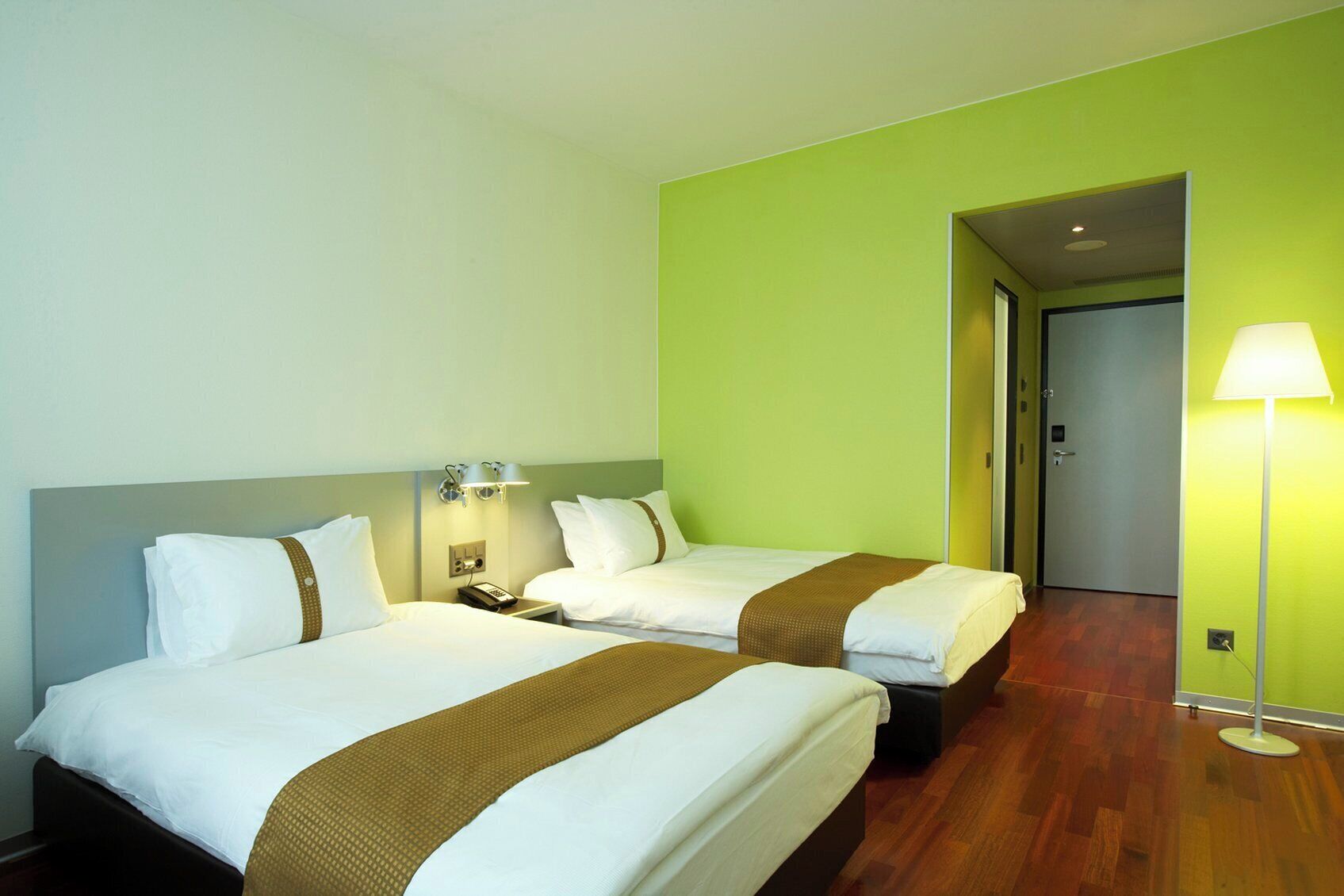 A hotel room with two beds, a nightstand, a lamp, and a door leading to another room.