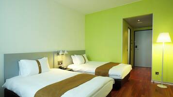 A hotel room with two beds, a nightstand, a lamp, and a door leading to another room.