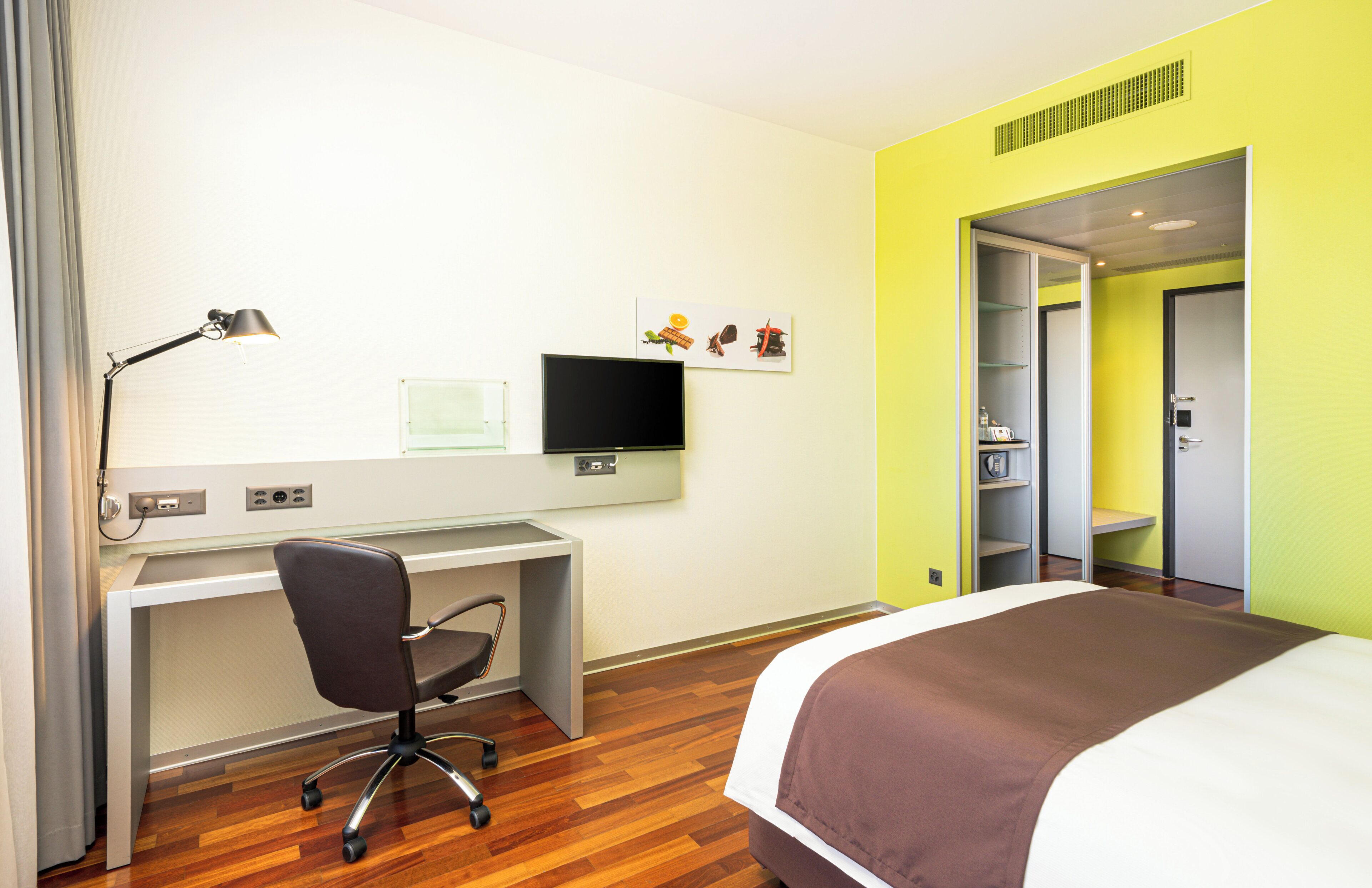 A hotel room with a bed, a desk with a chair, a television, and a wardrobe.