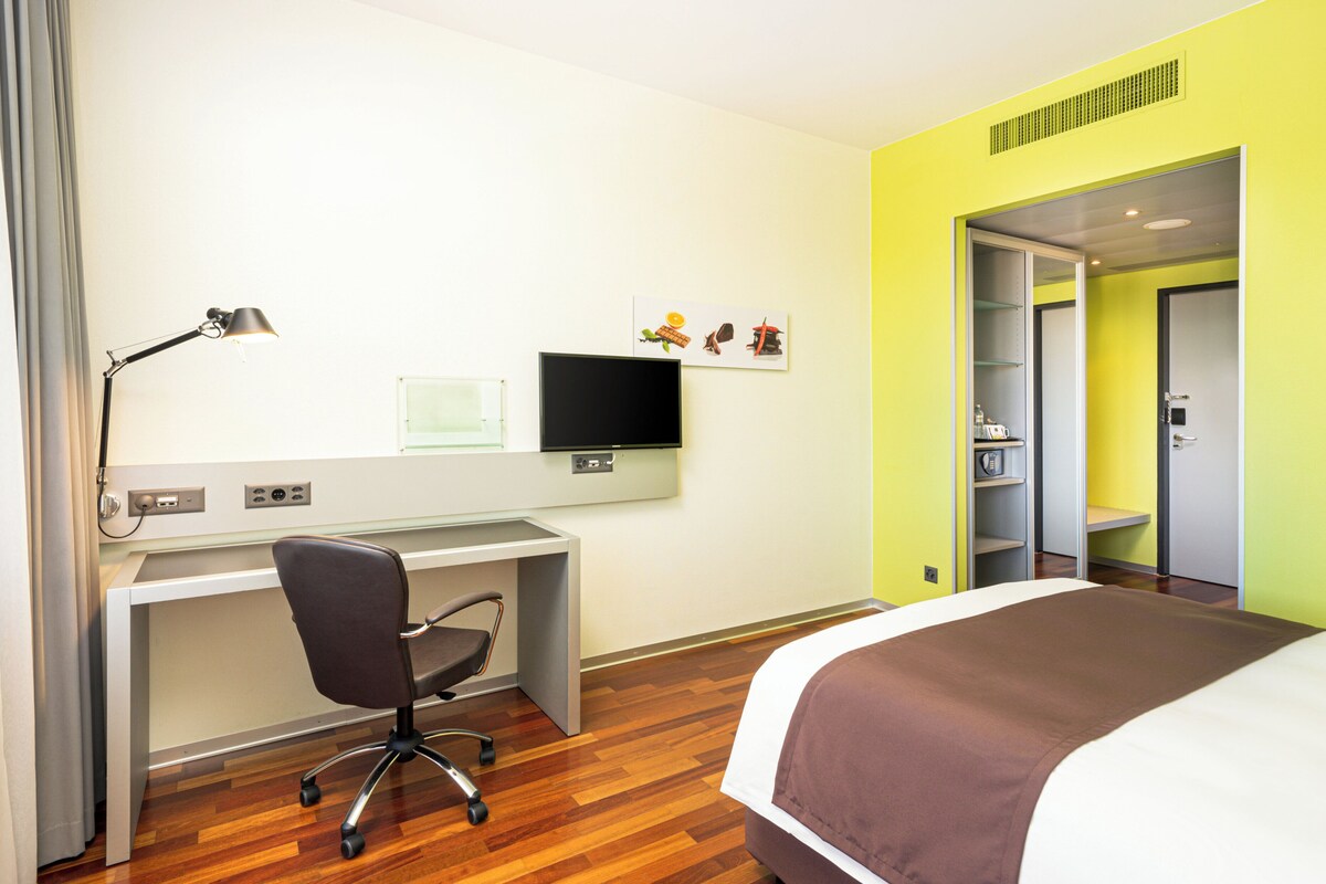 A hotel room with a bed, desk, chair, TV, and a door leading to a bathroom.