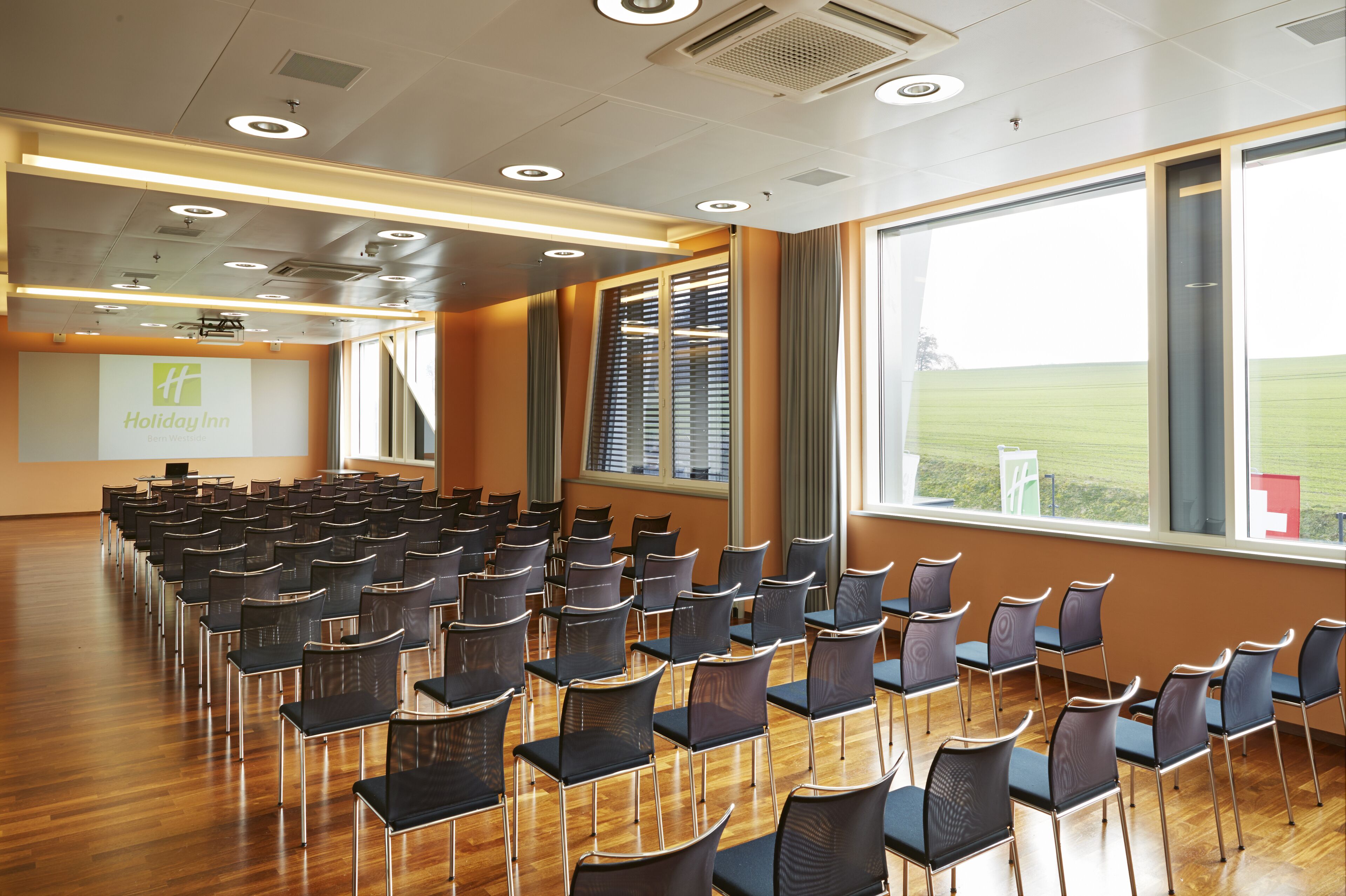 Un salón de conferencias con filas de sillas, una pantalla de proyector y grandes ventanas que dan a un campo.