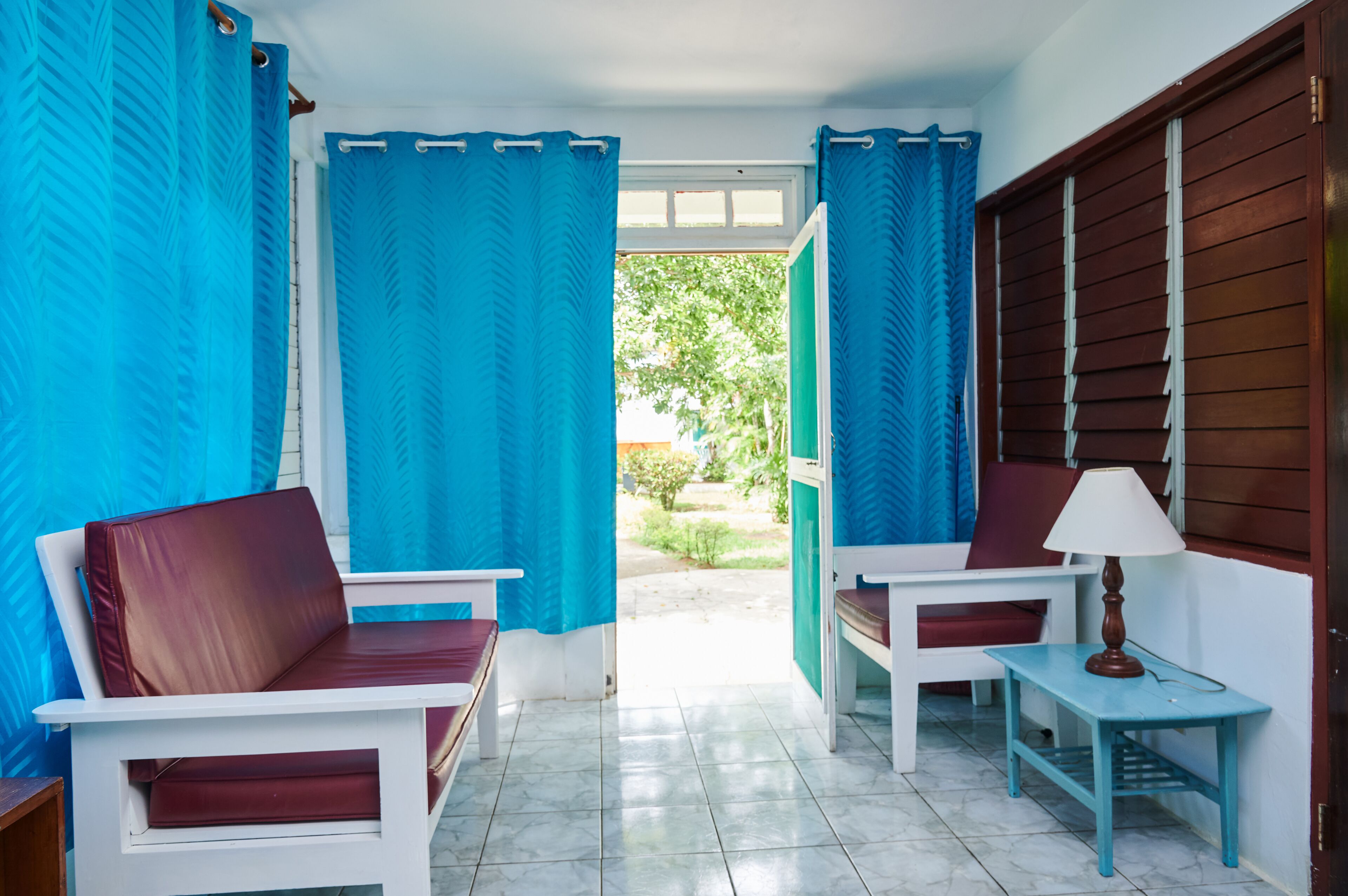 A room with two white chairs with red cushions, a small table with a lamp, and a view of a garden through blue curtains.