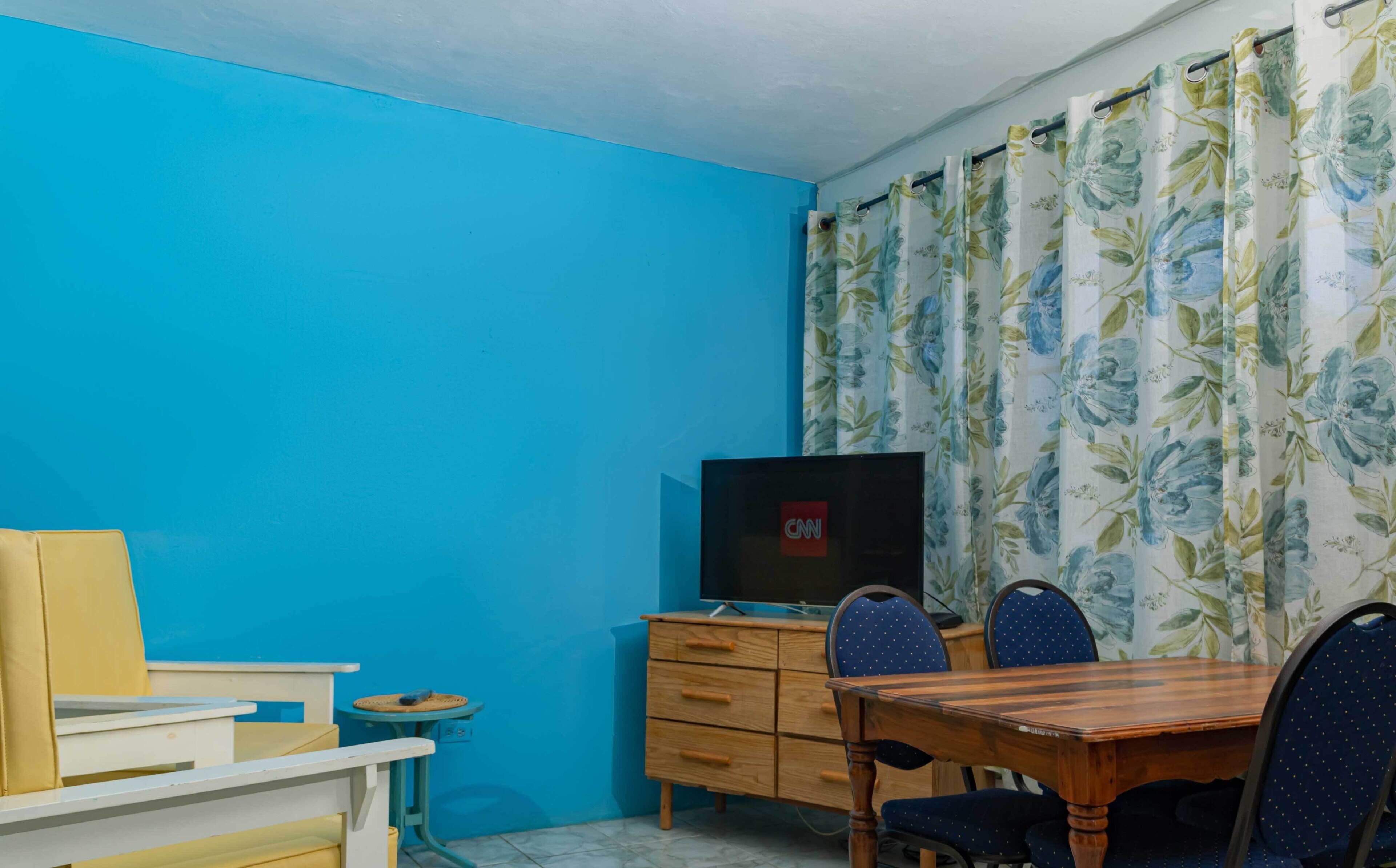 A room with a blue wall, a wooden table with chairs, a TV on a wooden cabinet, and floral curtains.