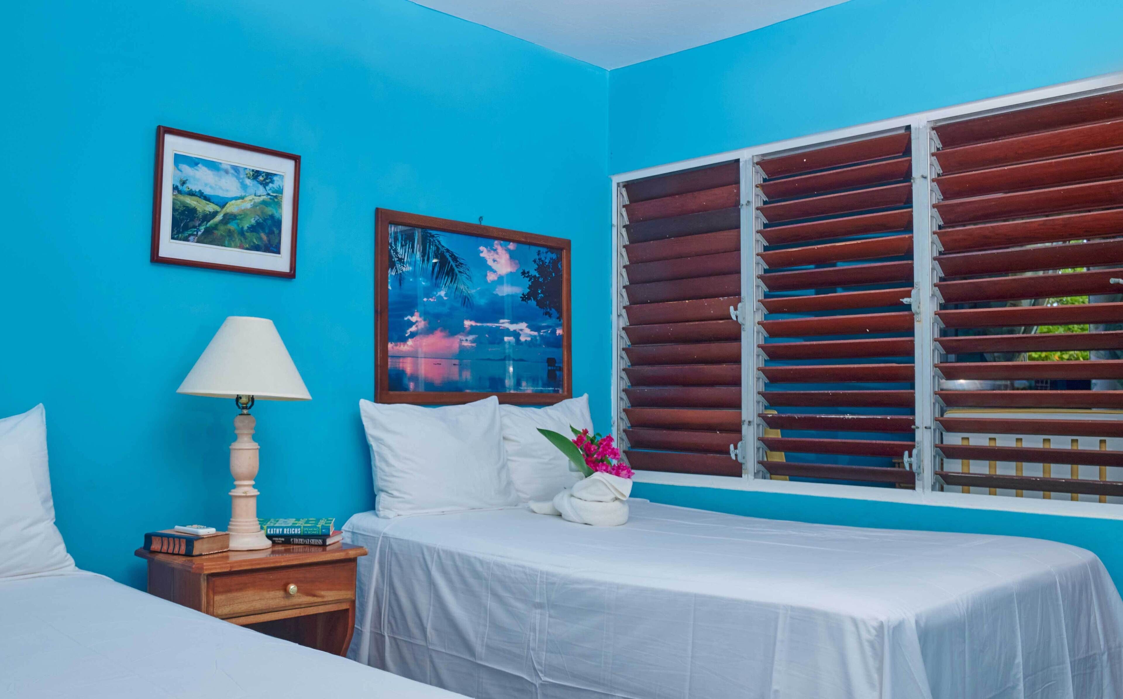 Una habitación con una cama, una mesita de noche con lámpara, un ventanal con persianas de madera y dos cuadros enmarcados en la pared.
