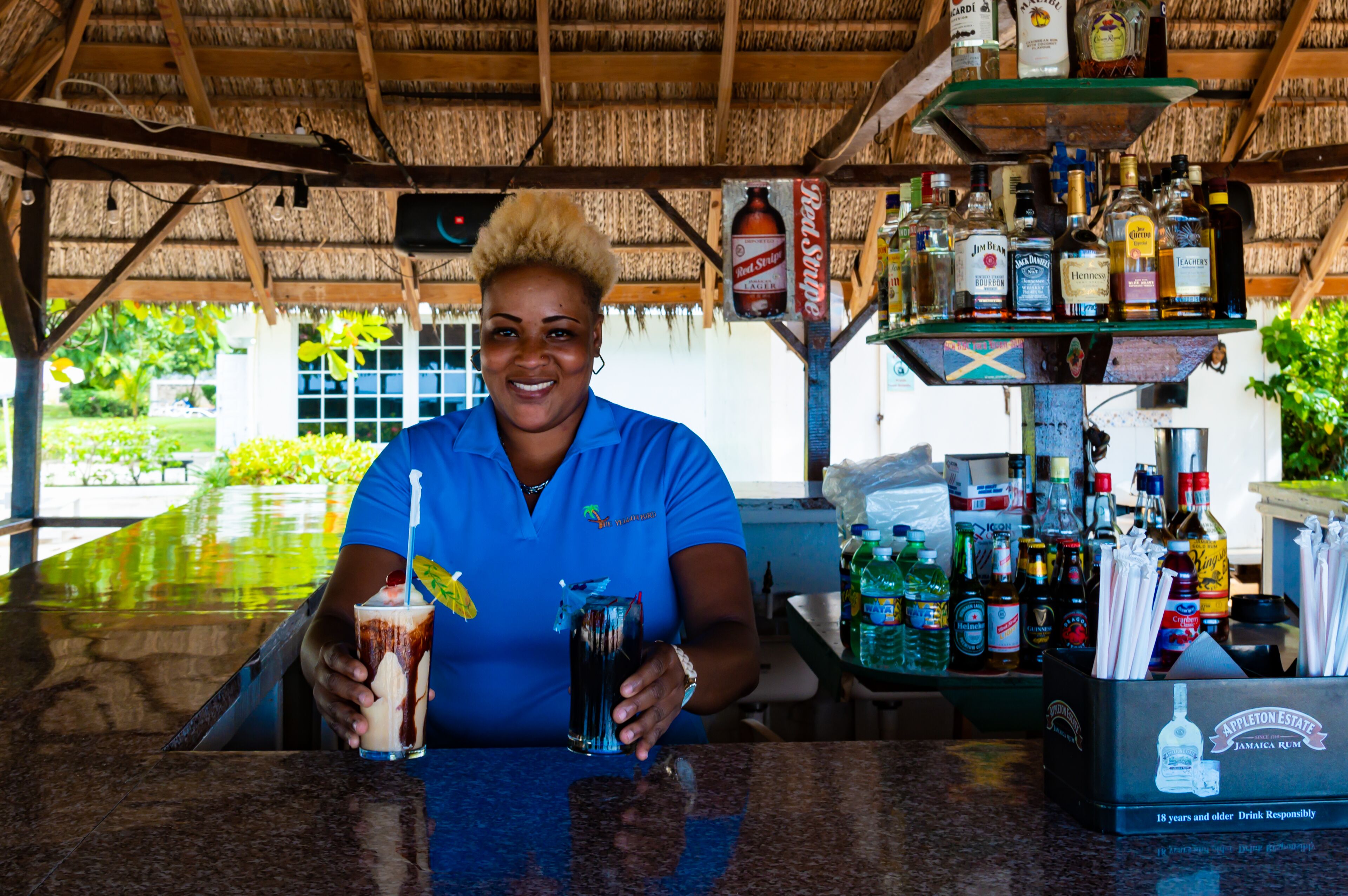 Une personne assise au bar, avec un verre devant elle, une variété de bouteilles d’alcool derrière, et un panneau indiquant l’âge légal pour consommer de l’alcool.