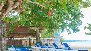 A beachside lounge area with blue and white lounge chairs under a tree, a wooden structure with a thatched roof, and a sign indicating beach rules. - The Yellow Bird (Negril)
