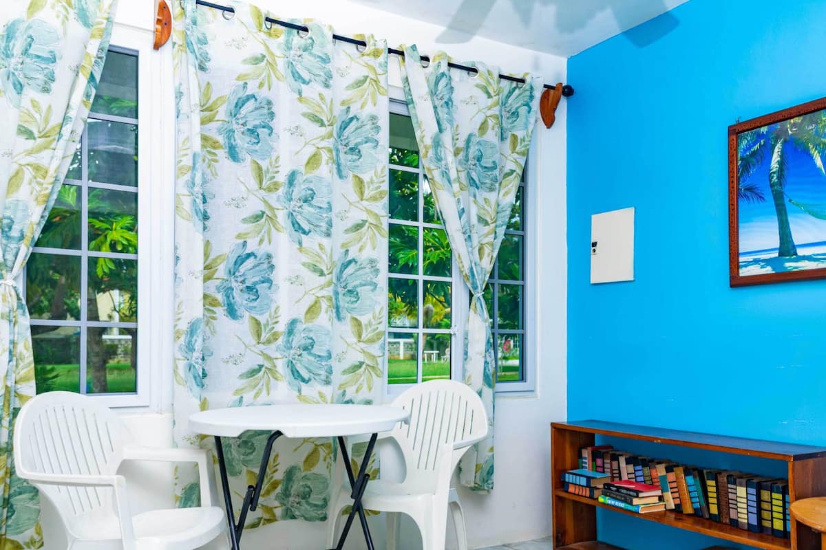 Une pièce avec un mur bleu, une fenêtre ornée de rideaux à motifs floraux, une table blanche avec deux chaises et une bibliothèque remplie de livres.