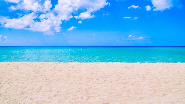 A beach with clear turquoise water and a sandy shore under a bright blue sky with scattered clouds. - The Yellow Bird (Negril)