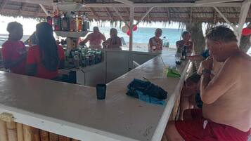 A beachside bar with a thatched roof, a counter with various bottles, and a view of the ocean.