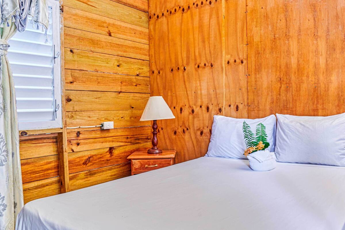 Une chambre à coucher avec une tête de lit en bois, une table de chevet avec une lampe et un lit recouvert de linge de lit blanc et agrémenté d’un coussin décoratif.