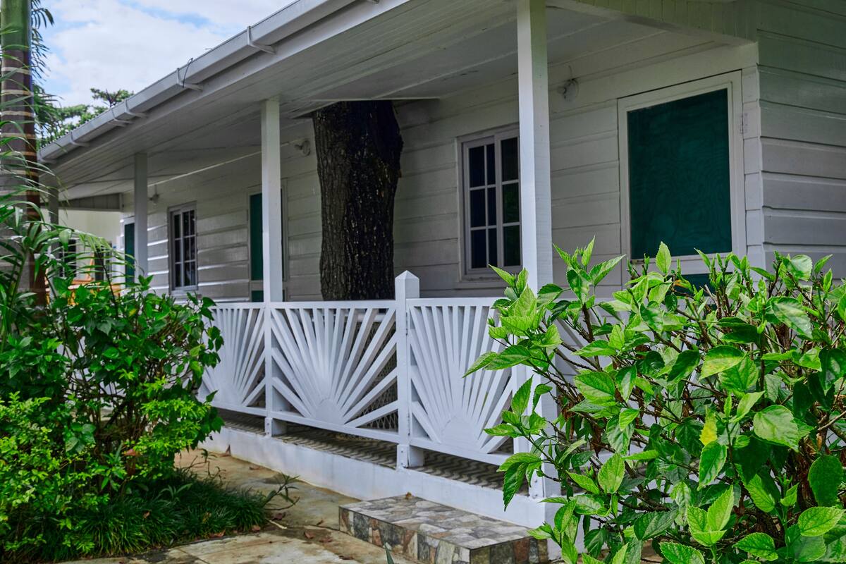 Une maison en bois blanche, avec un porche, des volets verts et un grand tronc d’arbre au centre.