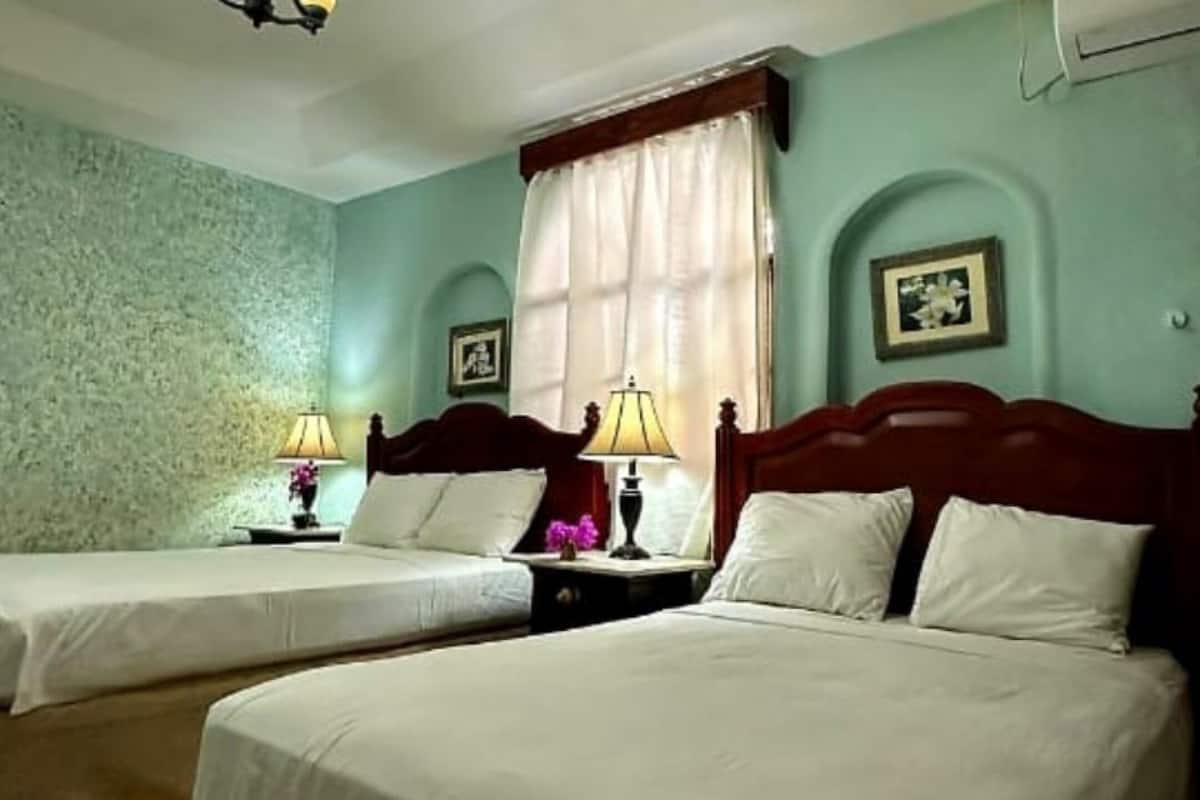 A room with two single beds, each with a wooden headboard, a nightstand with a lamp, and a window with a curtain.