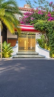Una casa con entrada protegida por un portón, camino de entrada con losetas y abundante vegetación, incluyendo una palmera y plantas con flores.