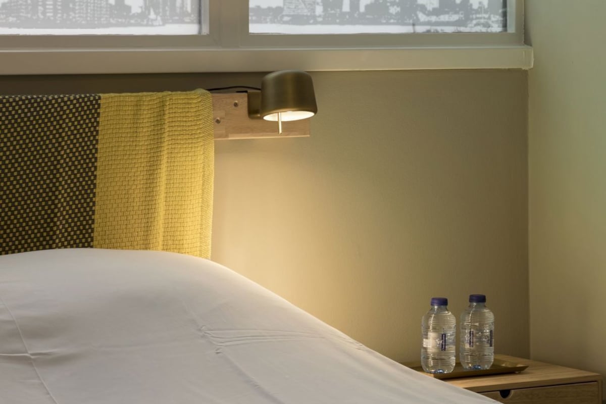 A hotel room with a bed, a window with a view of a city skyline, a lamp, and two water bottles on a nightstand.