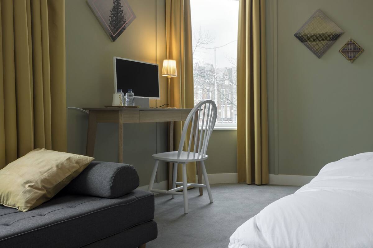 A hotel room with a bed, a desk with a computer, a chair, and a window with curtains.
