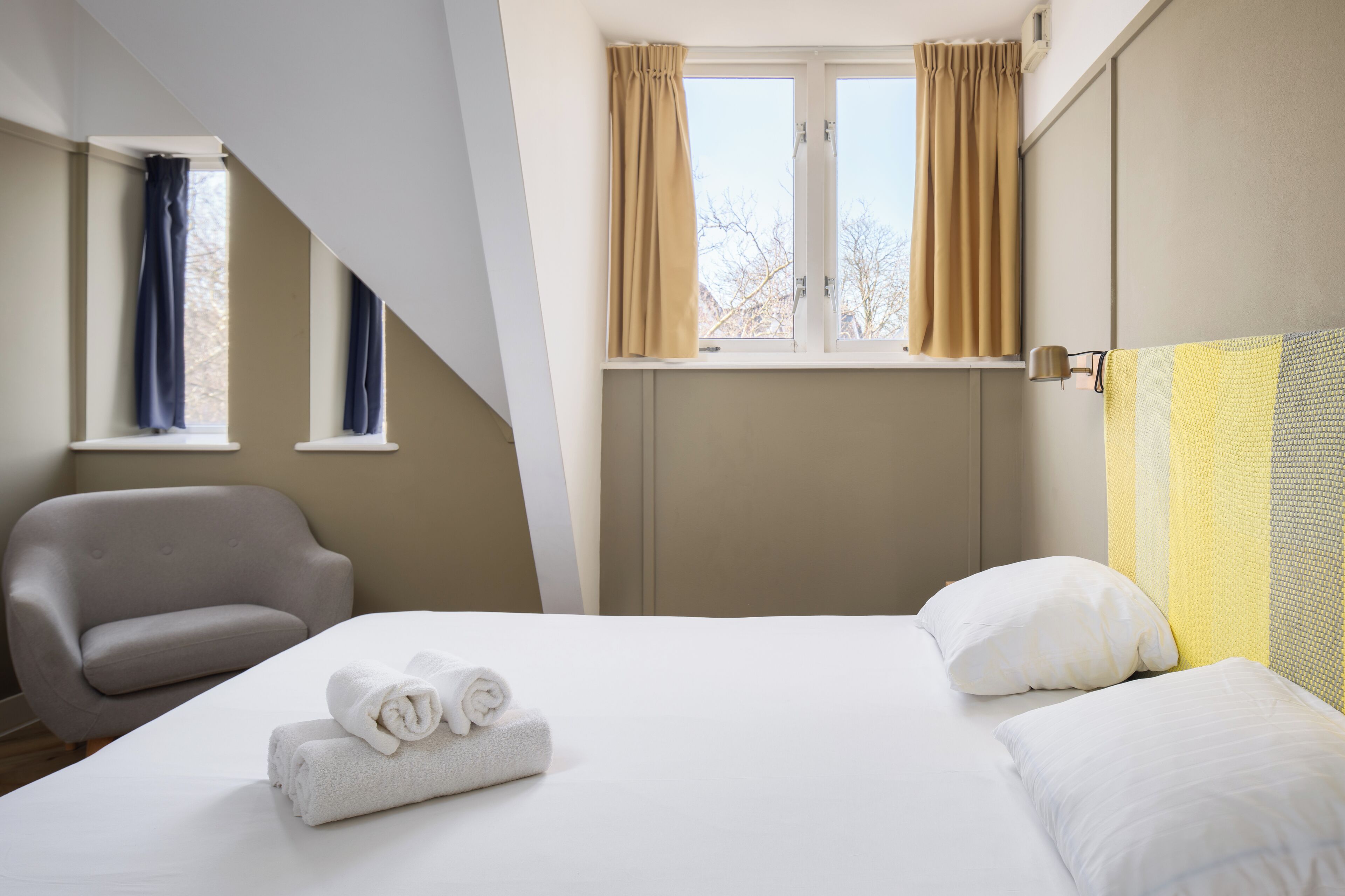 A hotel room with a bed, a chair, a window with curtains, and a view of trees outside.