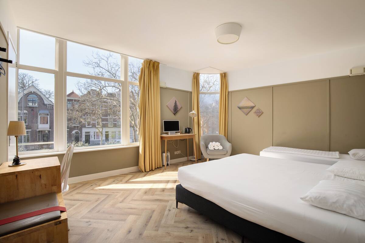 A hotel room with a large window, a desk with a computer, a chair, and a bed.