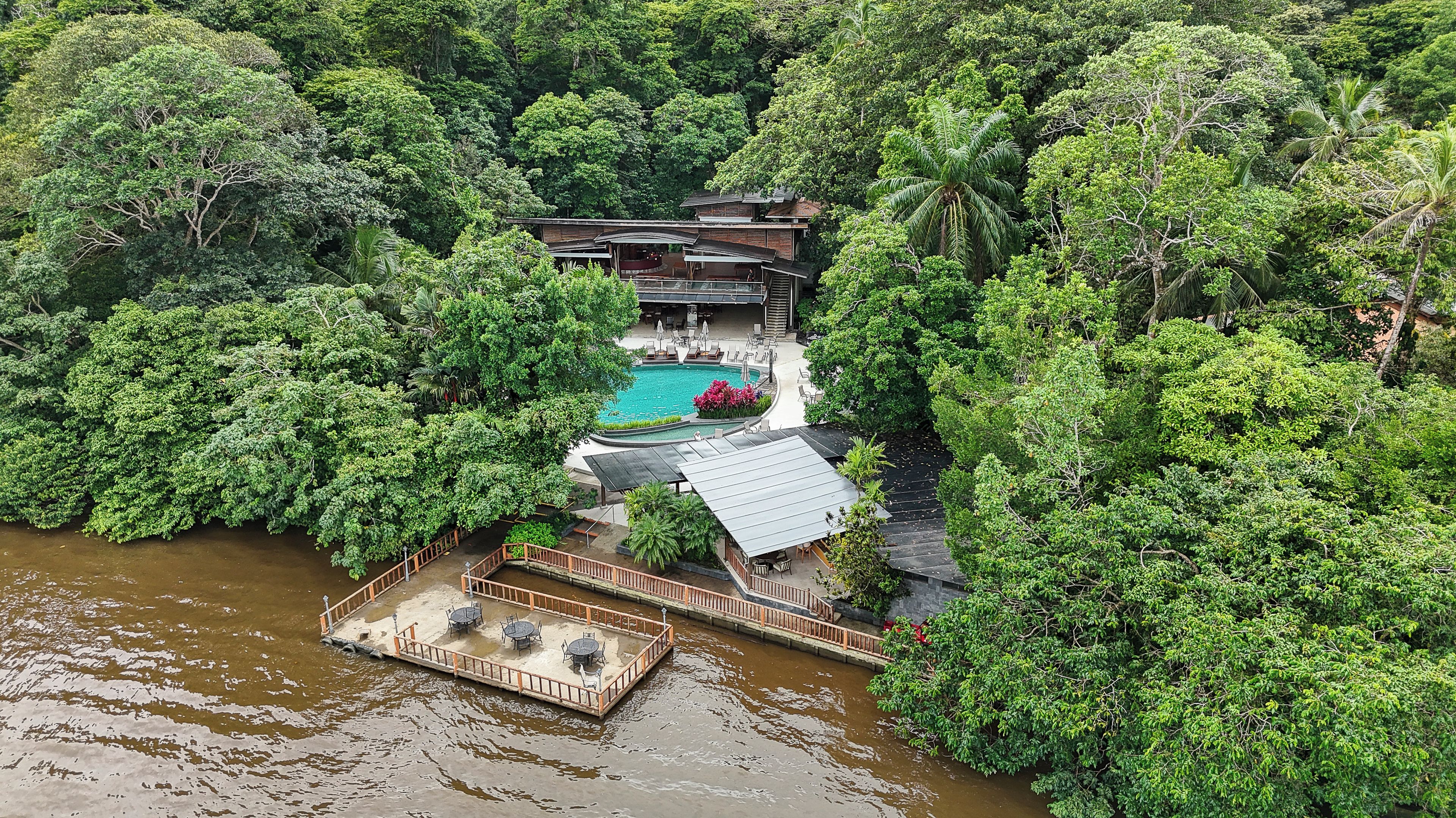 Un resort con piscina rodeada de exuberante bosque, un espacio de estar techado al aire libre y un muelle que se adentra en el agua.