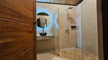 Un baño moderno con puerta de madera, un espejo redondo, un lavabo con cuenca de piedra, una ducha con cabezal de ducha manual y una toalla en el suelo.