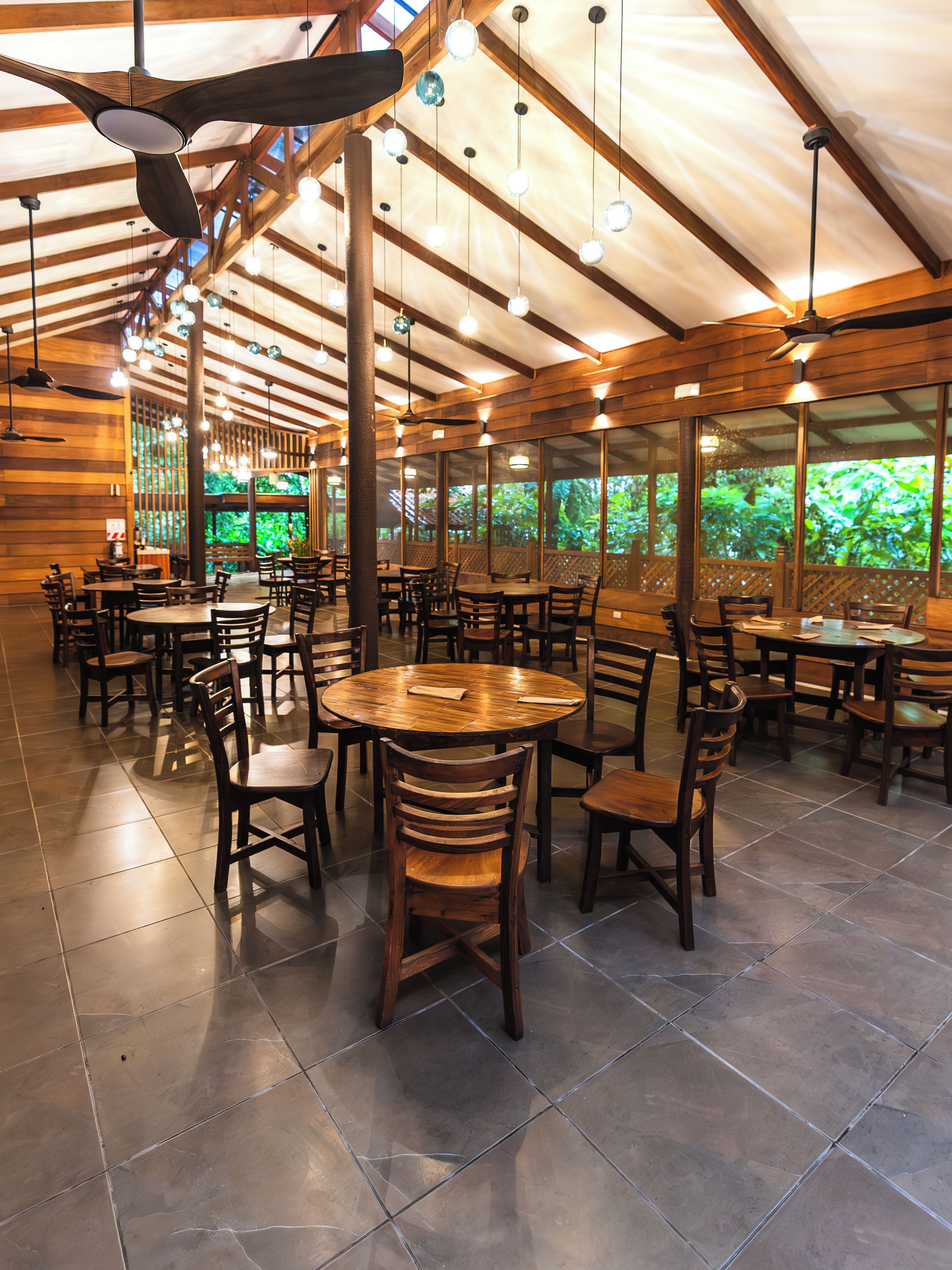 Un restaurant avec des tables et des chaises en bois, de grandes fenêtres et des lampes suspendues.