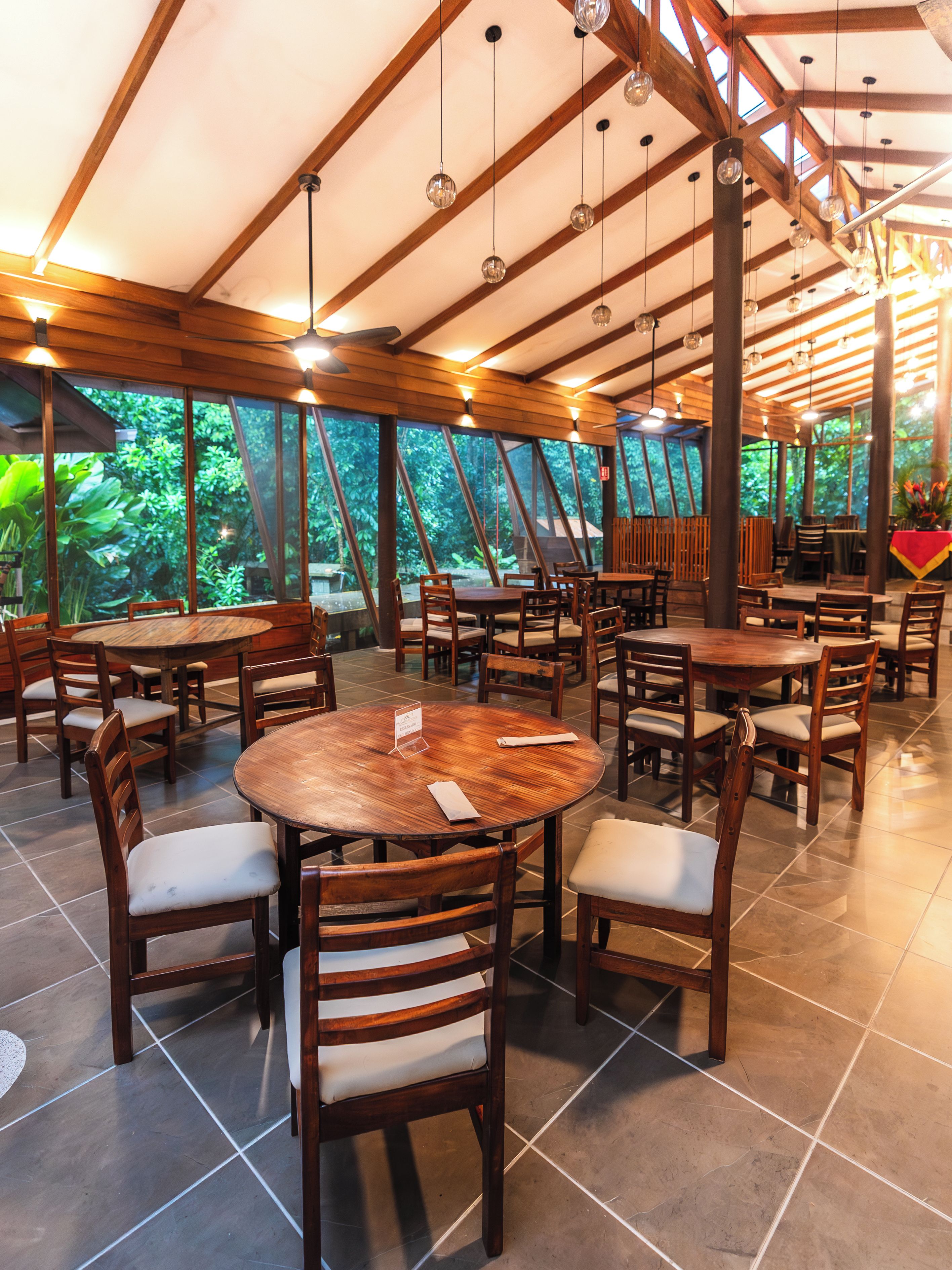 A restaurant with round tables and chairs, large windows, and a wooden ceiling structure.