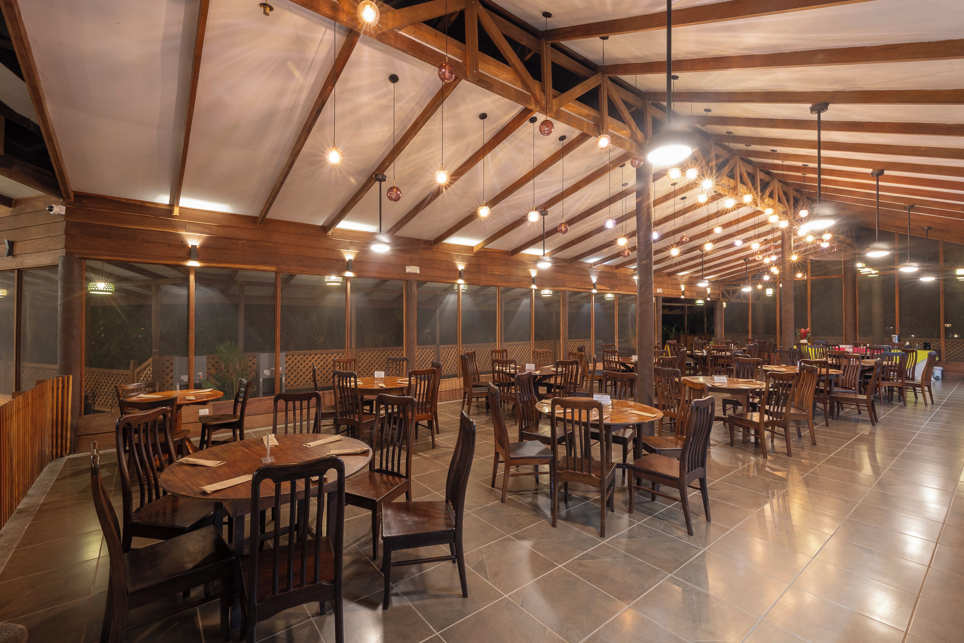 A spacious restaurant with high ceilings, wooden beams, and numerous hanging pendant lights. Tables and chairs are arranged for dining.