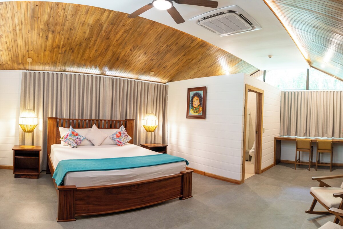 A bedroom with a wooden ceiling, a bed with a blue blanket, a desk with two chairs, and a painting on the wall.