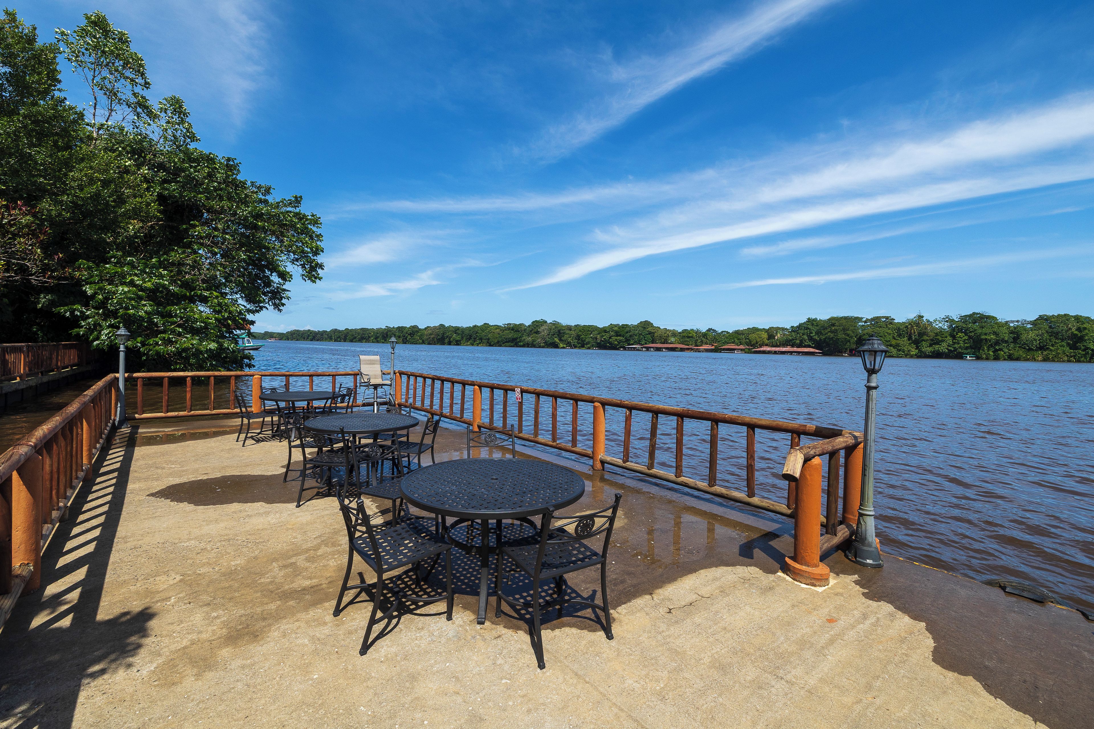 Eine Uferterrasse mit Tischen und Stühlen, einem Holzgeländer und Blick auf den See.