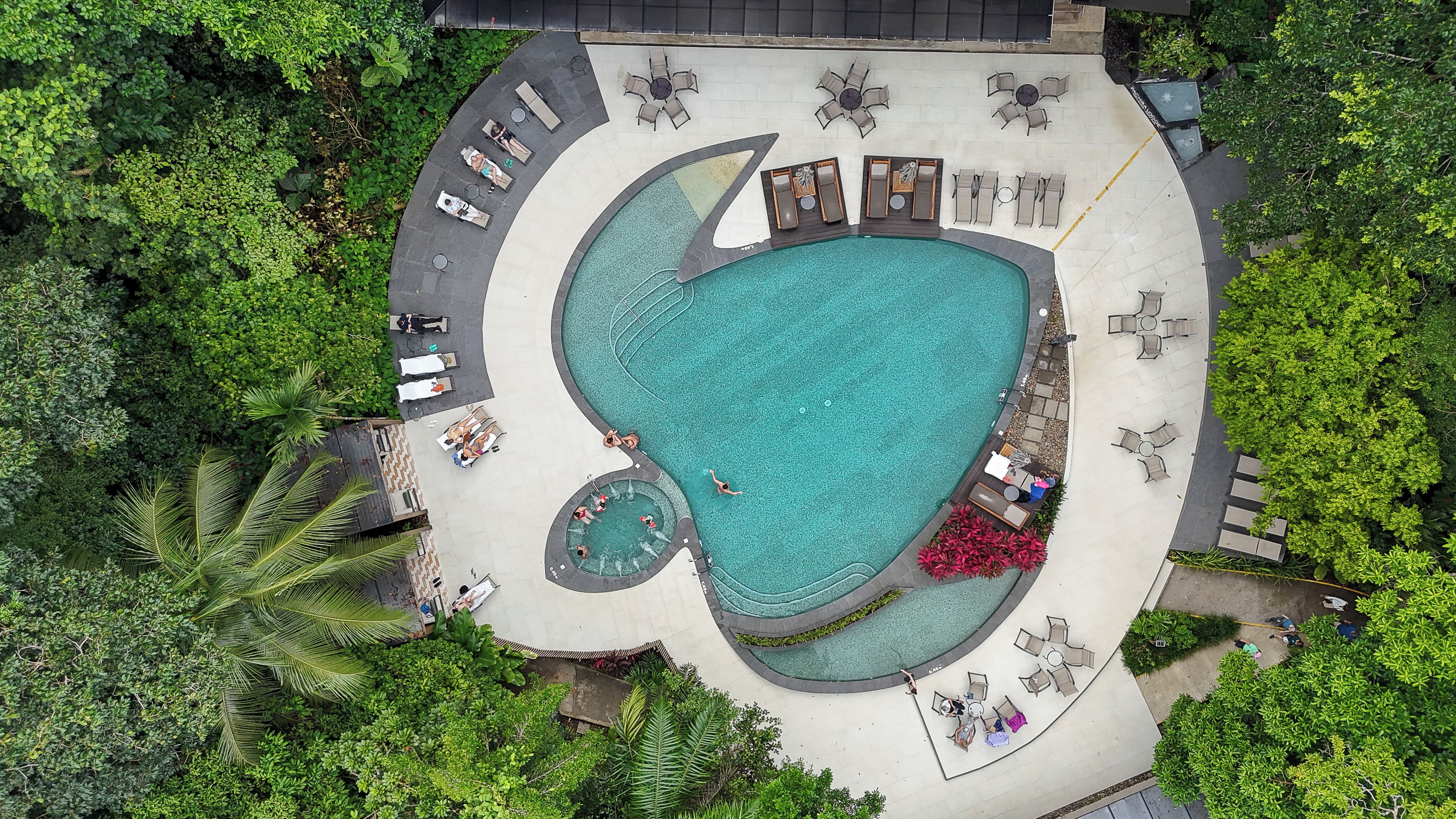 An aerial view of a resort with a large swimming pool, sun loungers, and surrounding greenery.