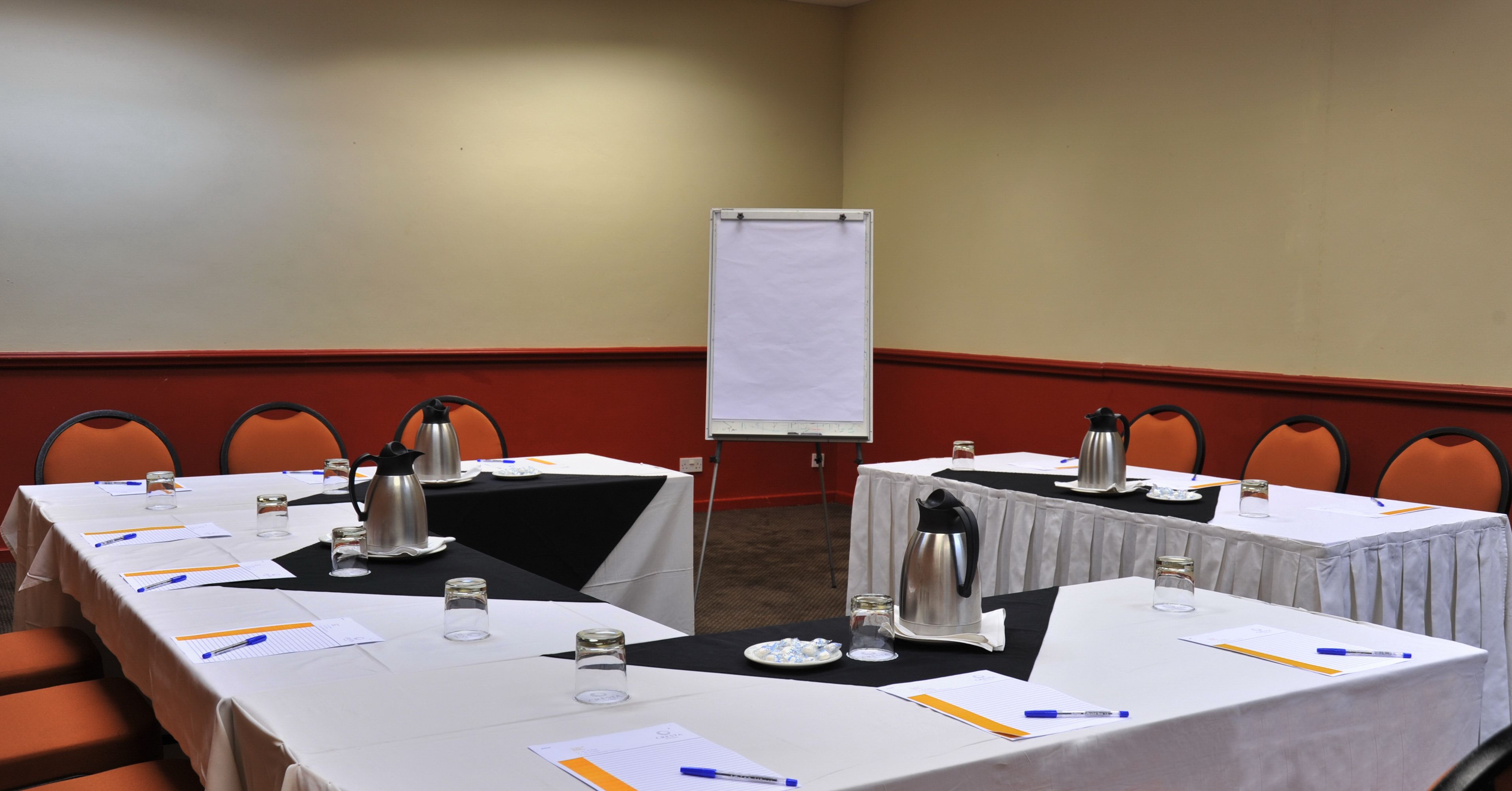 Un salón de conferencias con mesas dispuestas para reuniones, equipado con tarjetas de identificación, bolígrafos y cafeteras.