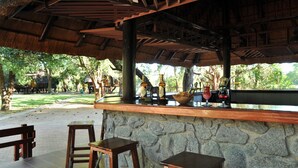 A covered outdoor bar area with stone walls, wooden stools, and a view of a natural landscape. - Cresta Marang Gardens (Francistown)