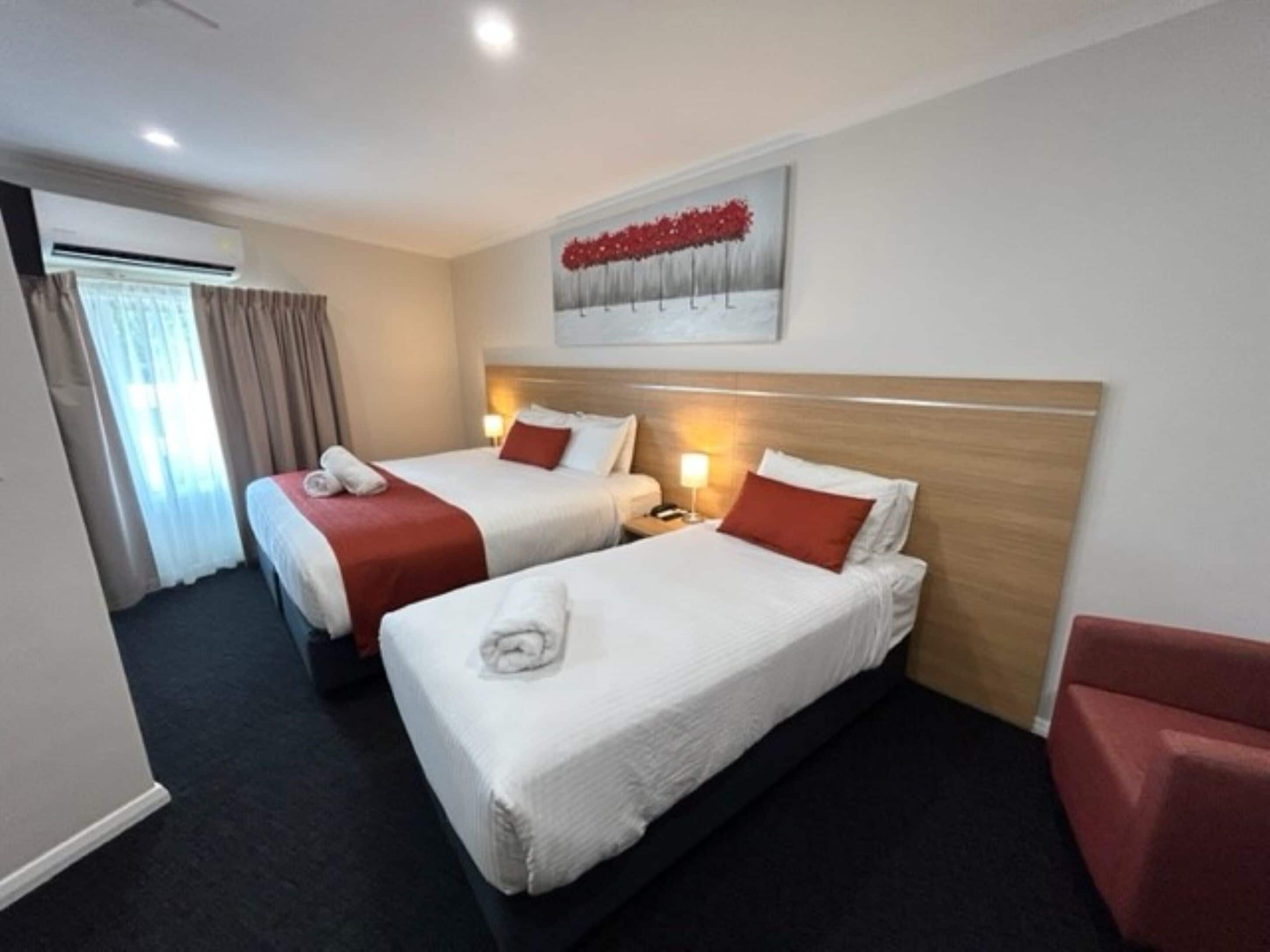 A hotel room with two beds, a red chair, a painting on the wall, and an air conditioning unit.