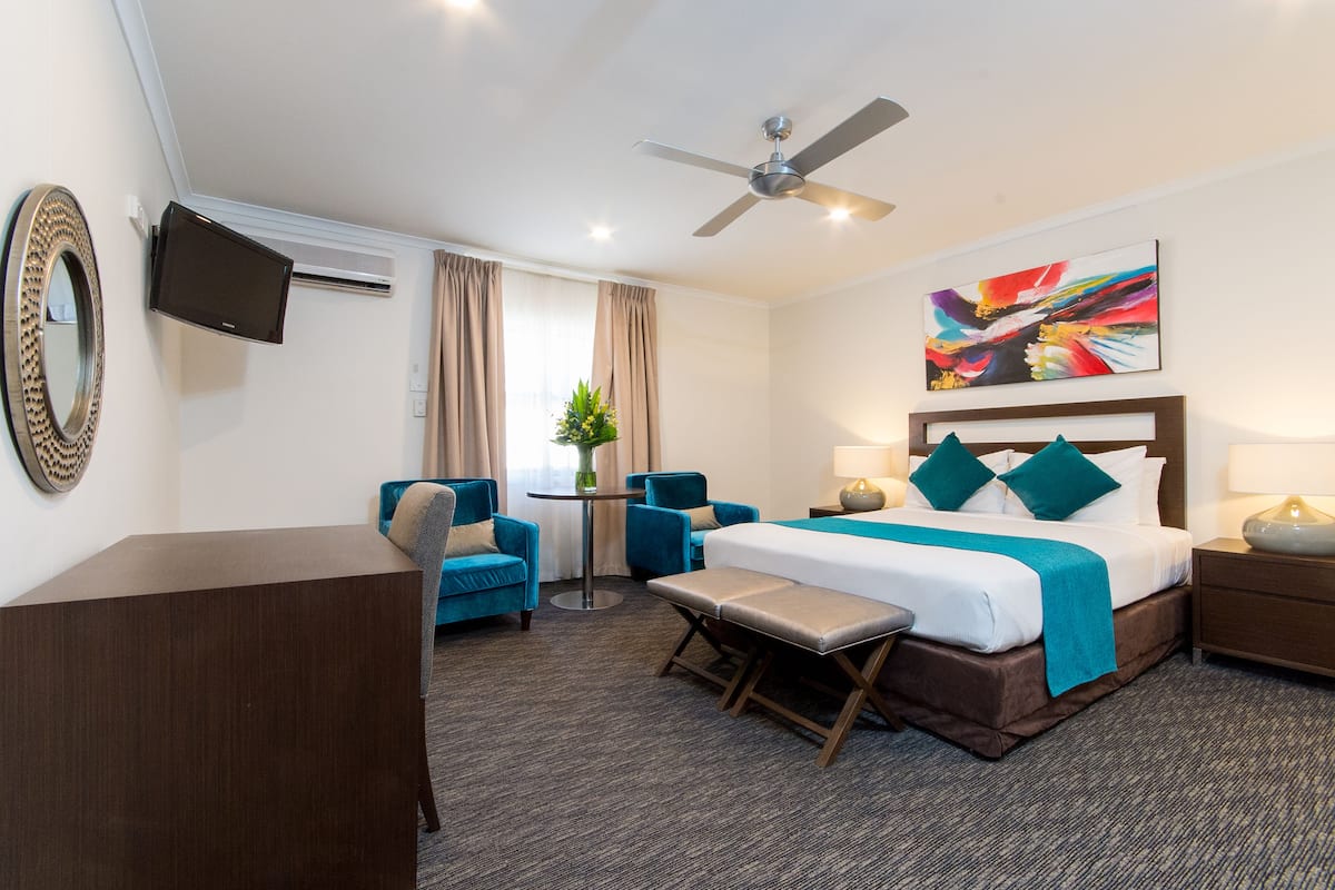 A hotel room with a bed, a desk, two chairs, a bench, a TV, a ceiling fan, a painting, and a window with curtains.