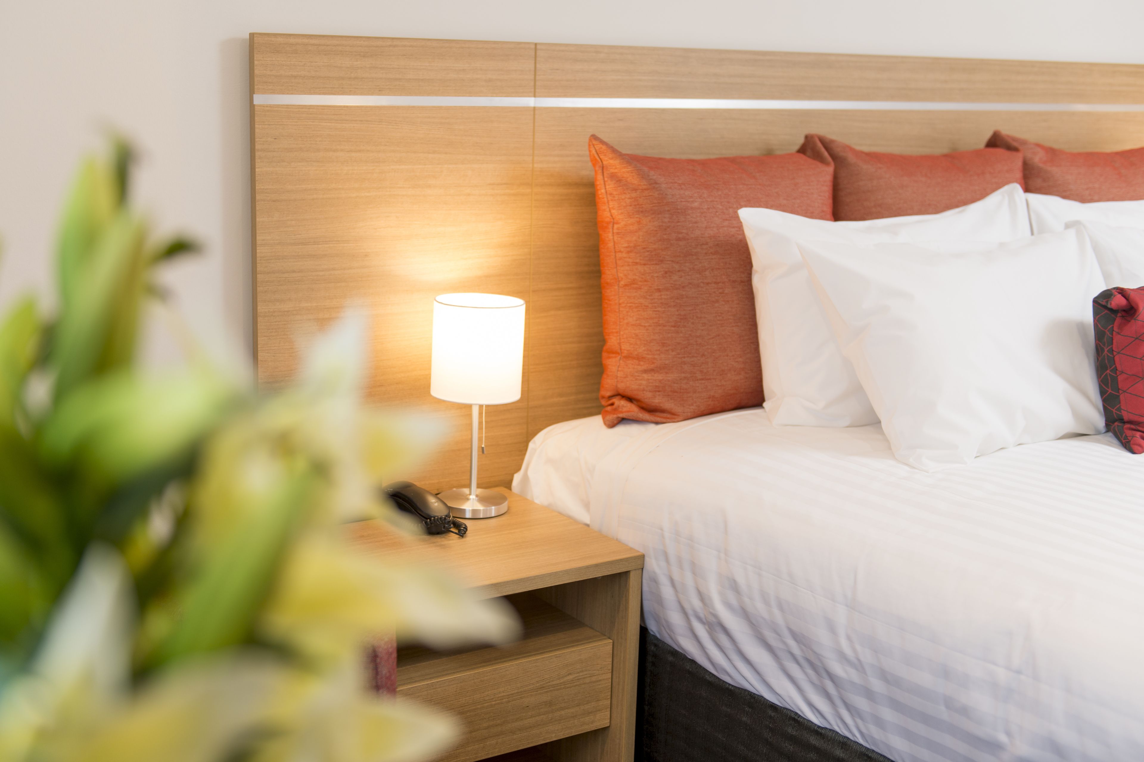 Una cama bien hecha con almohadas blancas y anaranjadas, una lámpara de noche y un cabecero de madera.
