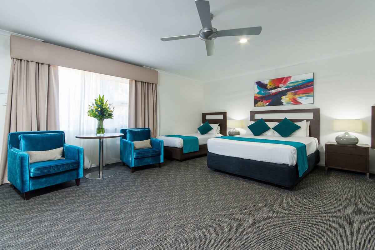 A hotel room with a large bed, two armchairs, a small table with a vase of flowers, a ceiling fan, and a painting on the wall.